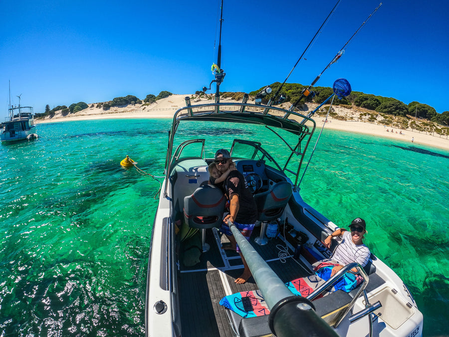 Two people on a boat in clear blue water with a beach and another boat in the background.